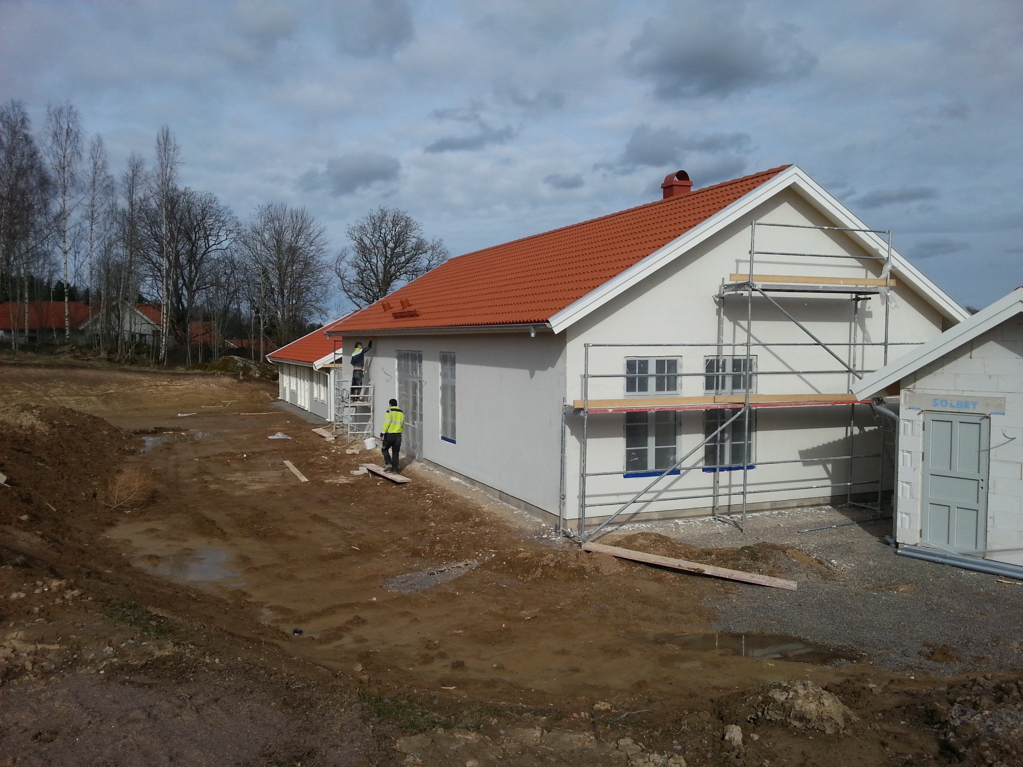 Biały dom z czerwoną dachówką w trakcie prac wykończeniowych. Widoczne rusztowania, pracownik w żółtej kamizelce i teren wokół domu w stanie surowym.