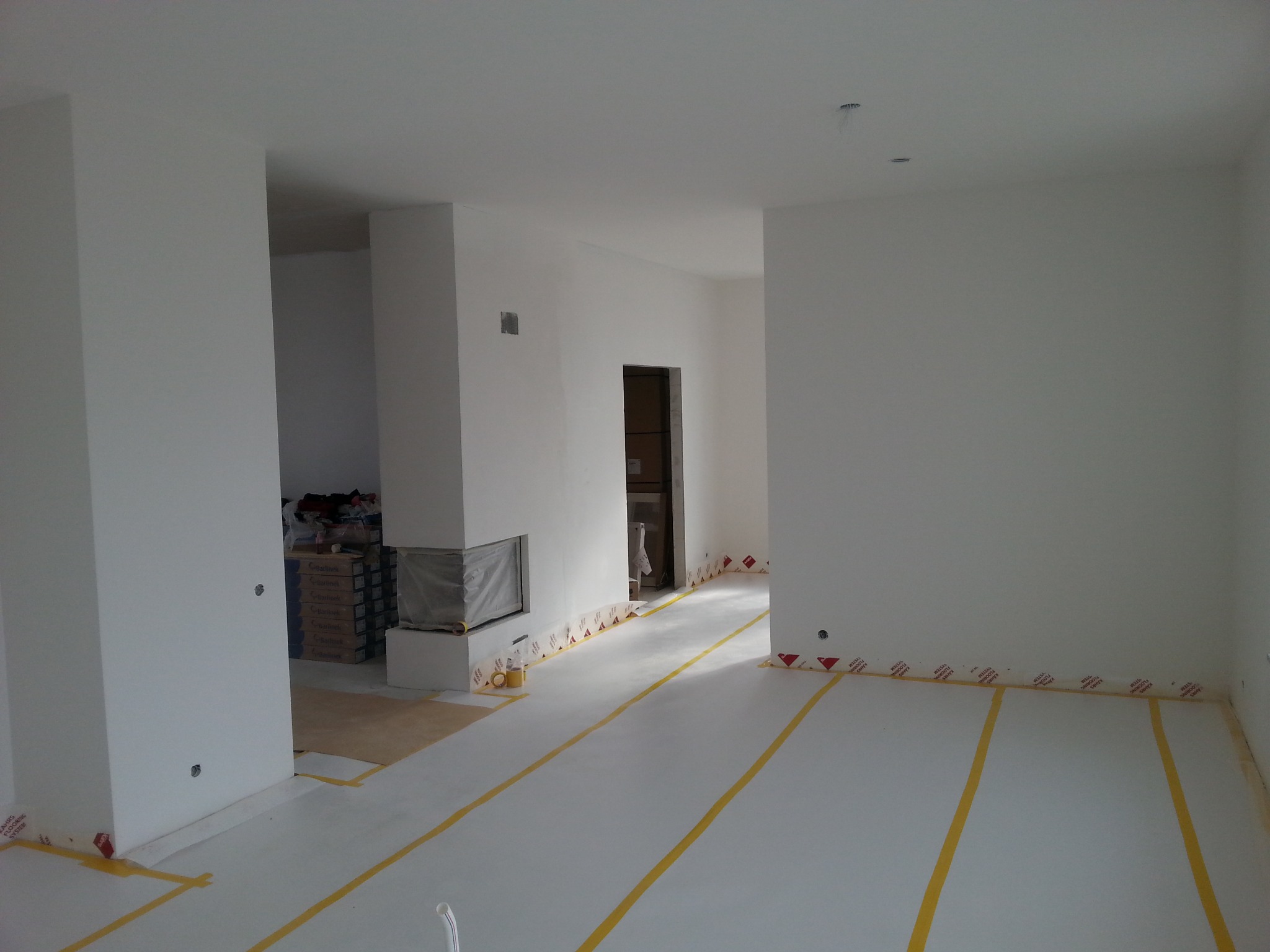 Wnętrze po szpachlowaniu i malowaniu na biało, z zabezpieczoną podłogą taśmą. Widoczne przygotowane materiały i fragment kominka. Minimalistyczny design w trakcie wykończenia.