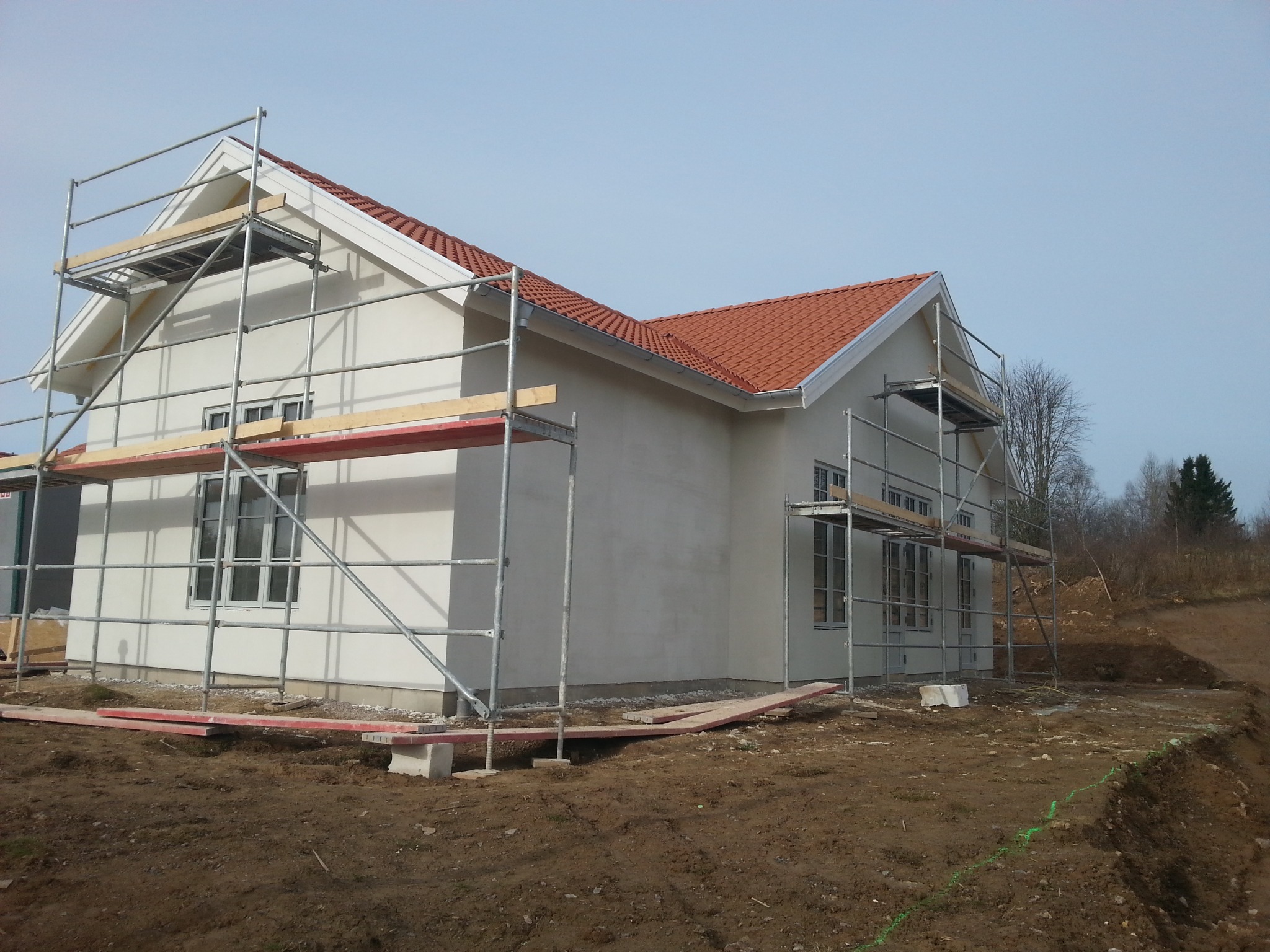 Biały dom z czerwoną dachówką w trakcie budowy, otoczony rusztowaniami. Widoczne okna i przygotowany teren wokół budynku.