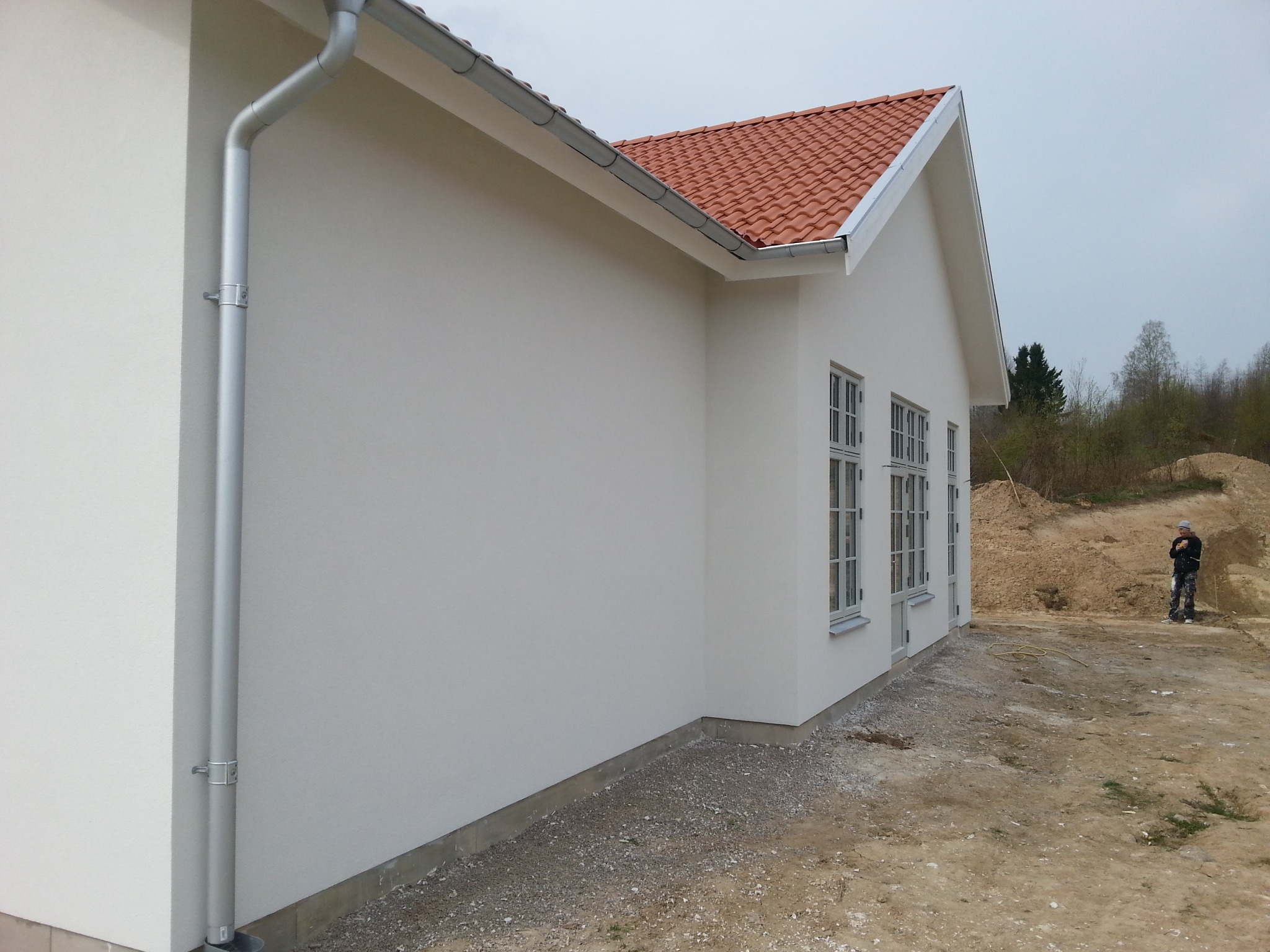 Boczny widok otynkowanego domu z czerwoną dachówką i szarymi rynnami. Na zewnątrz widoczna osoba w oddali i teren przygotowany pod zagospodarowanie.