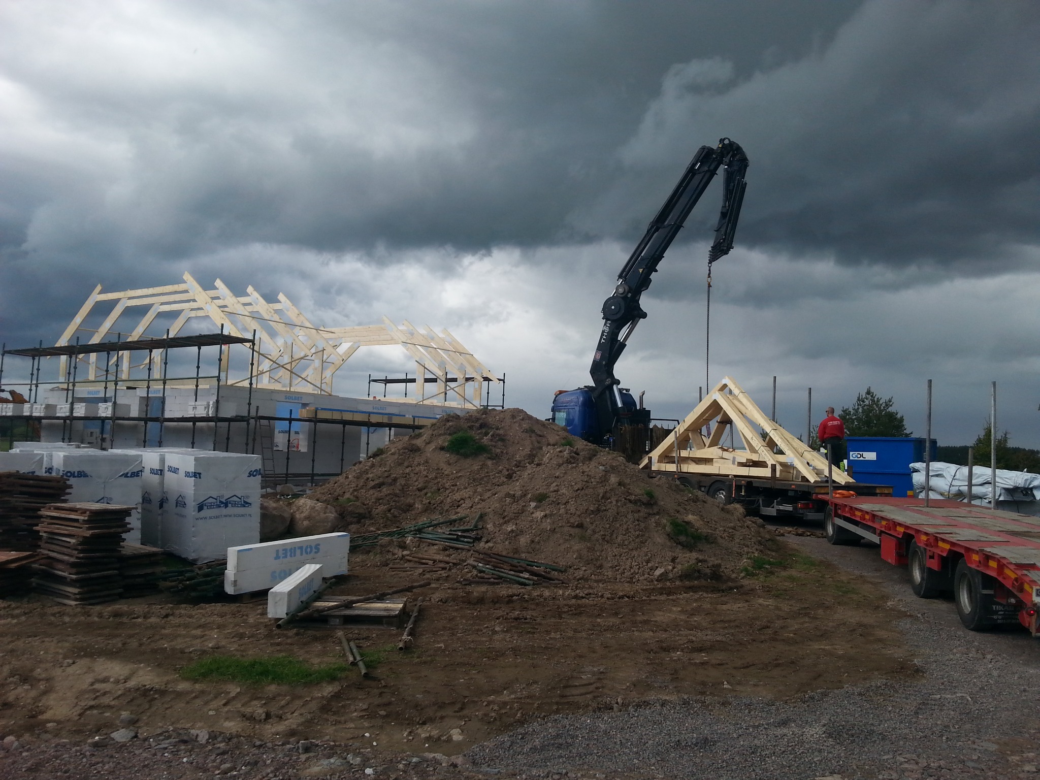 Montaż drewnianej konstrukcji domu szkieletowego za pomocą dźwigu na tle pochmurnego nieba. Widoczne materiały budowlane i elementy konstrukcyjne.