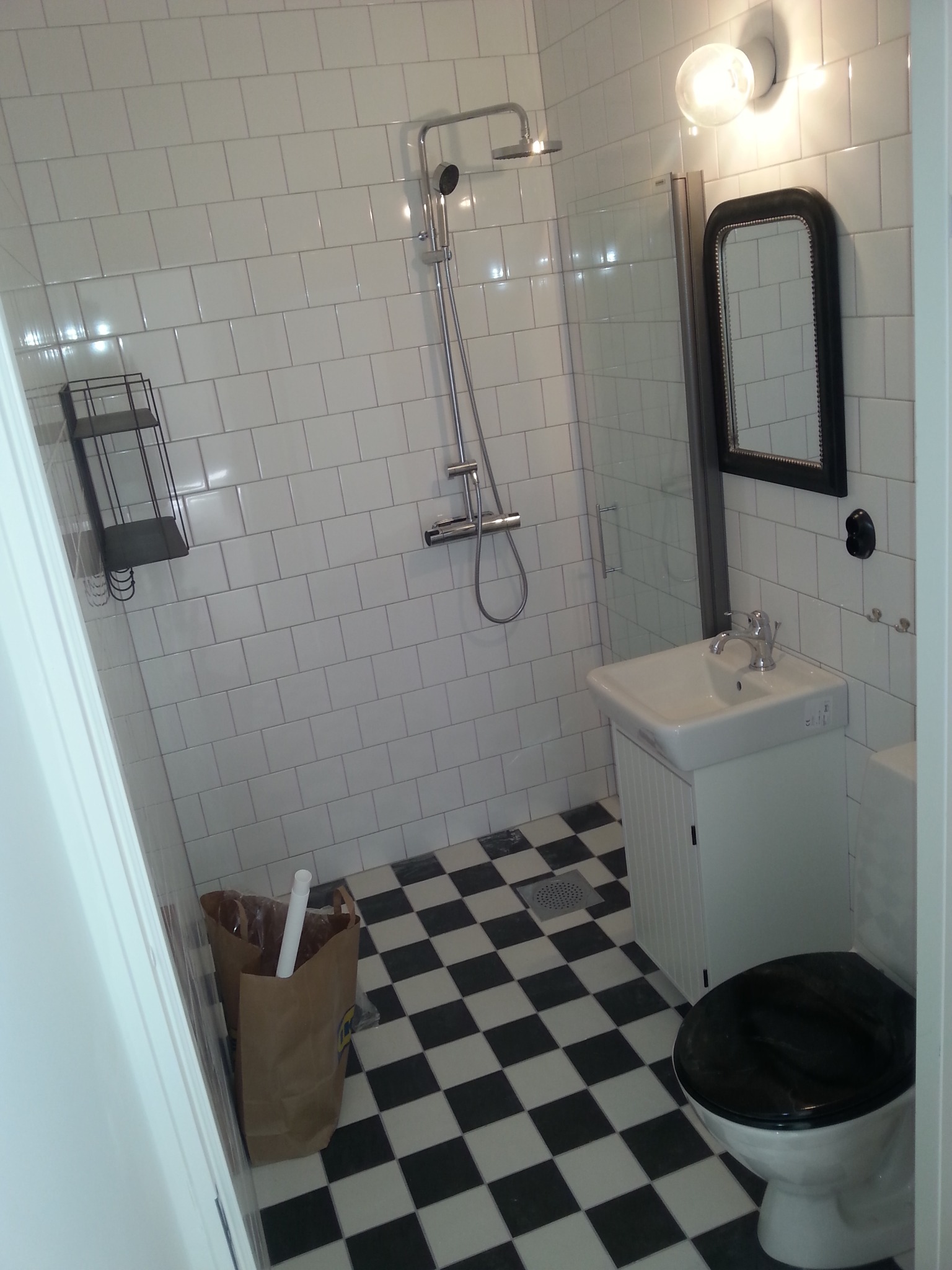 Wykończona łazienka z białą armaturą, kabiną prysznicową i czarno-białą podłogą w szachownicę. Na ścianach białe, prostokątne płytki.