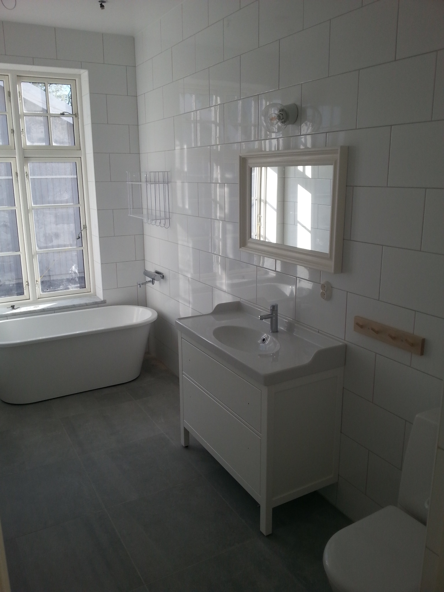 Jasna łazienka z wanną wolnostojącą, białą umywalką z szafką i dużymi, białymi płytkami na ścianach. Minimalistyczny design z szarymi płytkami podłogowymi.