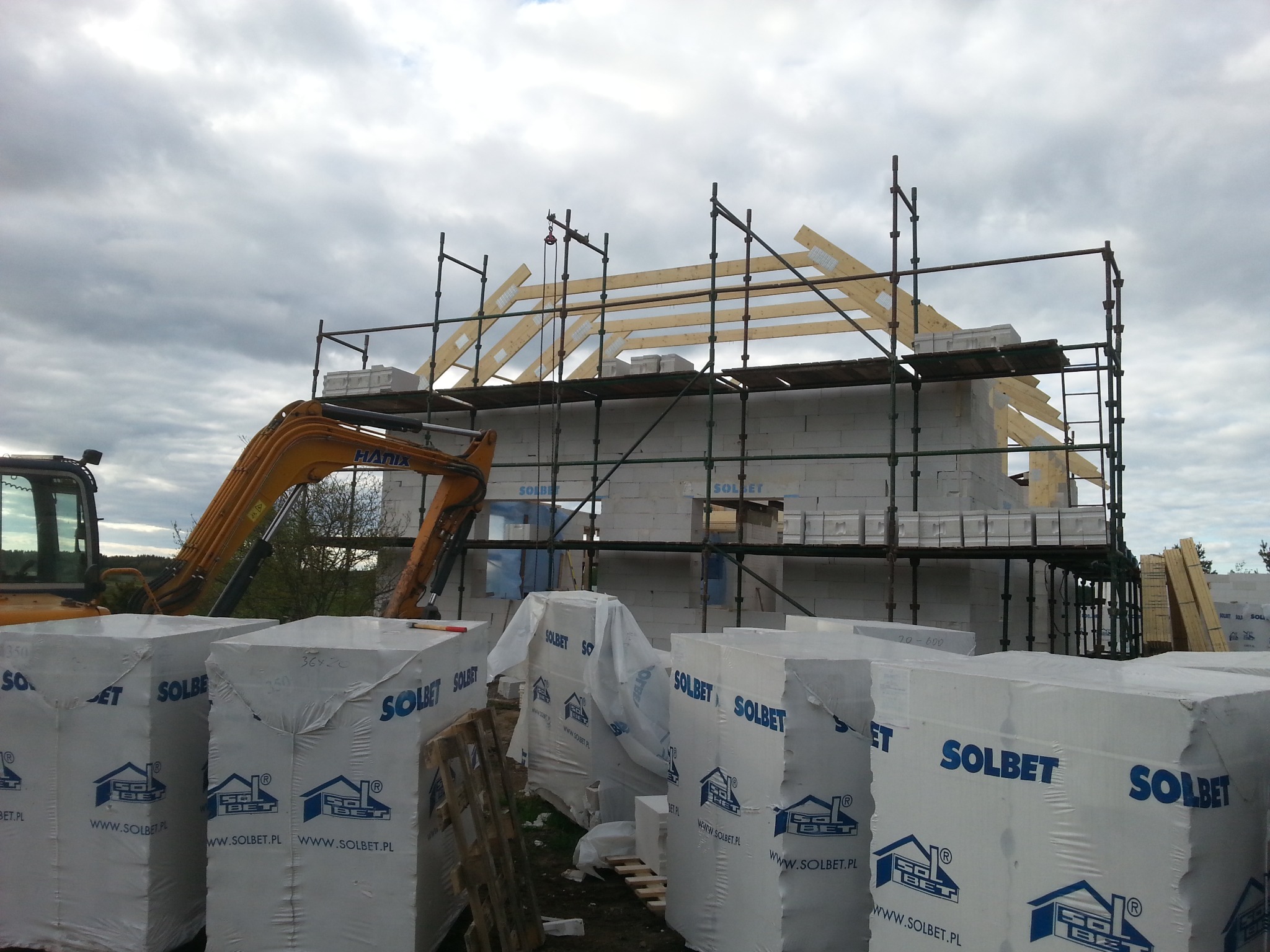 Budowa domu z bloczków Solbet, widoczne rusztowania, dźwig i drewniana konstrukcja dachu na tle pochmurnego nieba. Materiały budowlane na pierwszym planie.