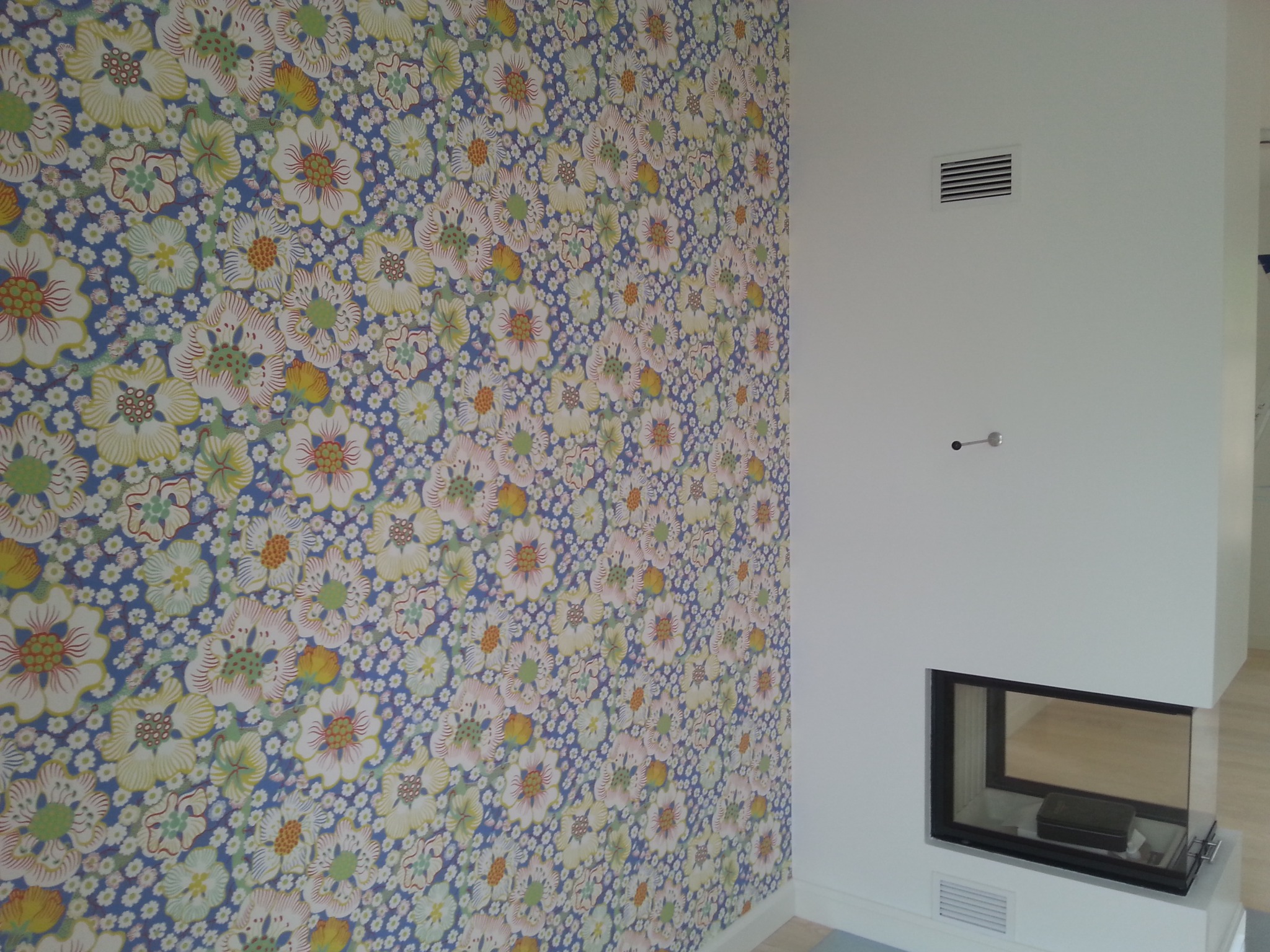 Ściana z tapetą w kwiaty i biały kominek z szybą. Widoczna kratka wentylacyjna i fragment podłogi. Wykończenie wnętrza w jasnych barwach.