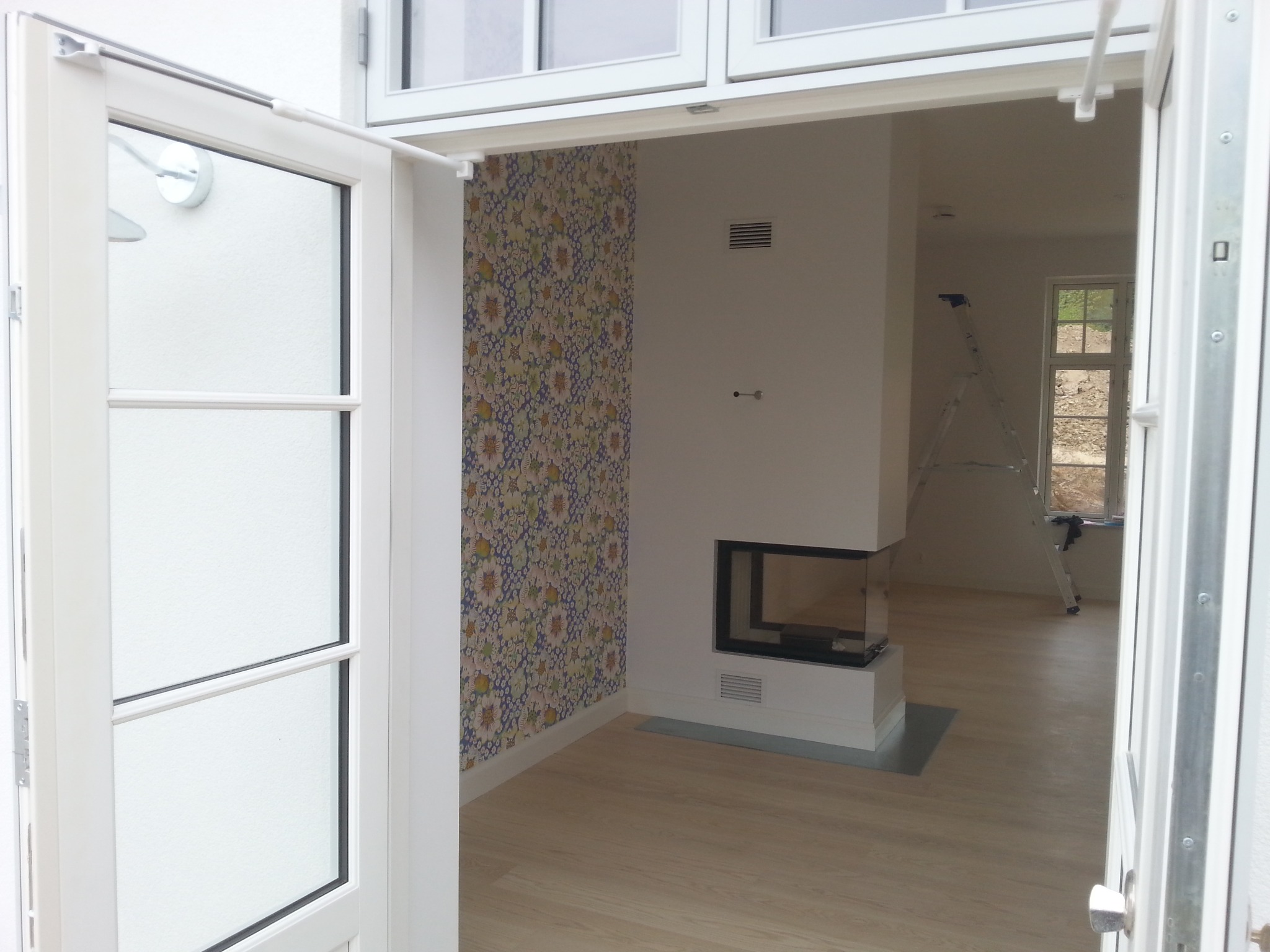 Otwarty widok na jasne wnętrze z kominkiem i wzorzystą tapetą, widziane przez białe, uchylone drzwi balkonowe. W tle drabina oparta o ścianę.