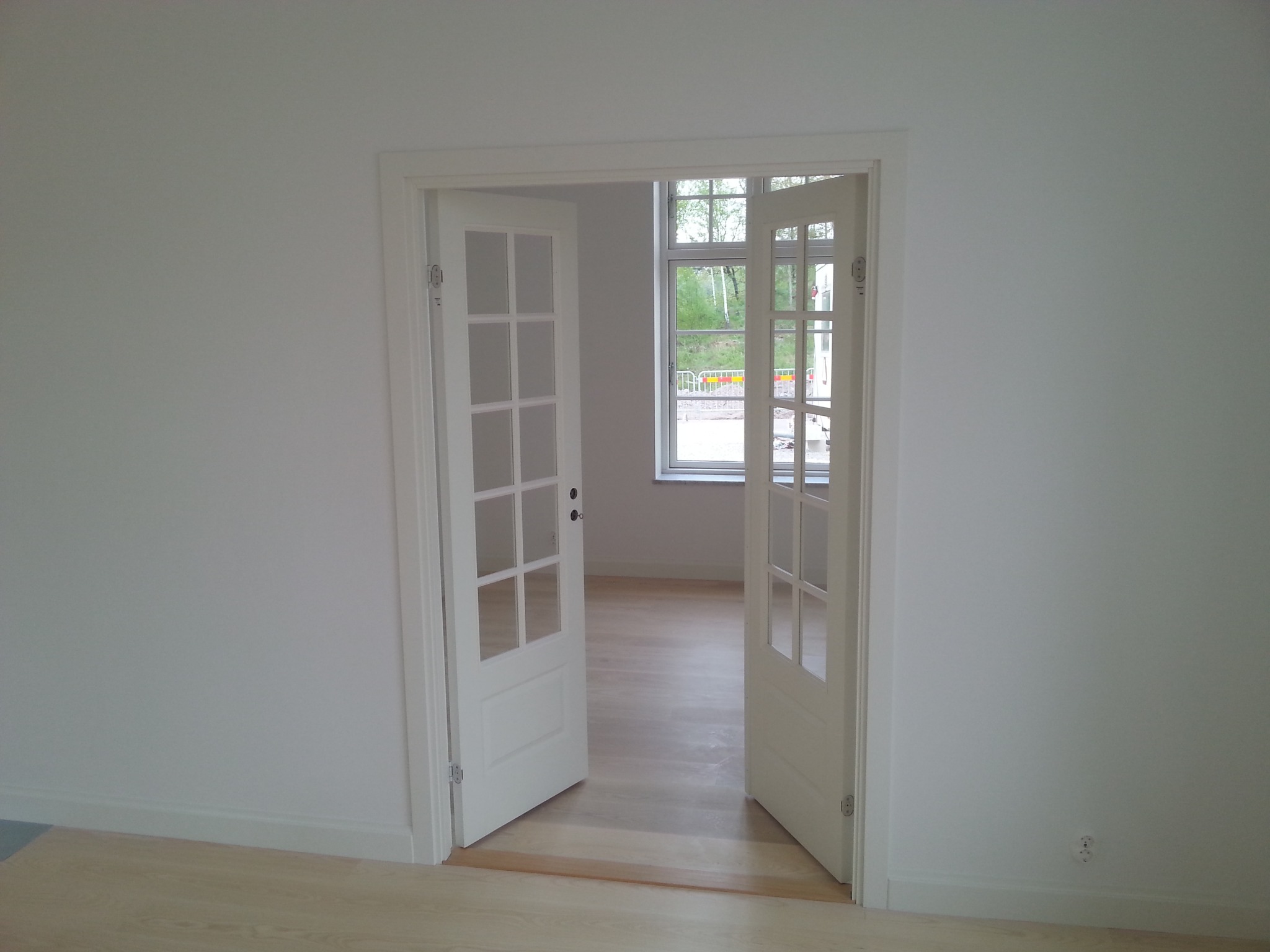 Białe, przeszklone drzwi dwuskrzydłowe, otwarte do wnętrza jasnego pokoju z drewnianą podłogą. Widok na okno z zielenią za nim. Minimalistyczne wykończenie.