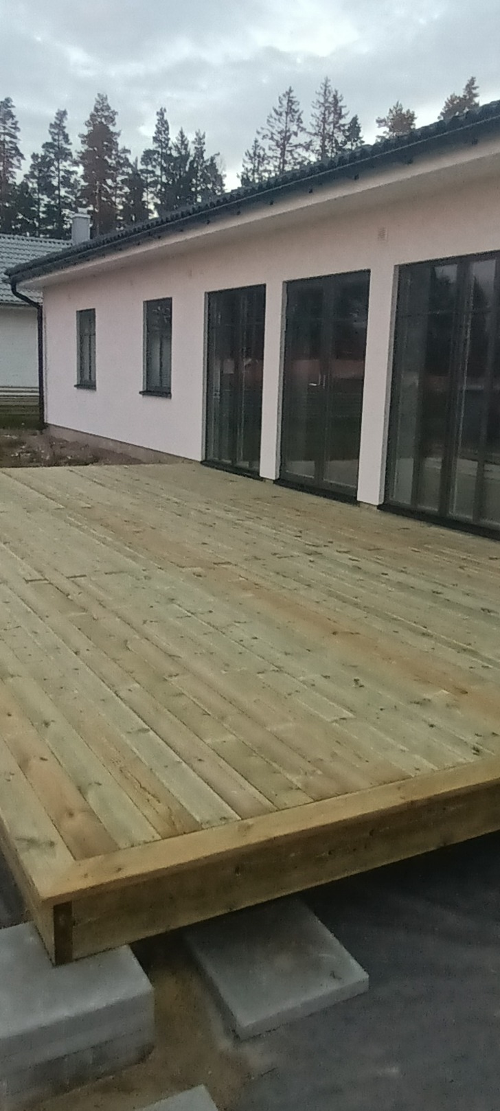 Nowo wybudowany drewniany taras przylegający do białego domu z dużymi oknami, widok na szare niebo i drzewa w tle.