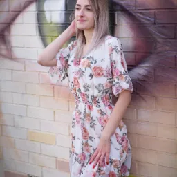 Kobieta w kwiecistej sukience pozuje na tle ceglanej ściany z muralem oka. Subtelny uśmiech, naturalne światło, swobodna poza. Sesja w plenerze, artystyczne tło.