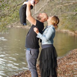 Fotodawka - Rodzina nad brzegiem jeziora: ojciec podnosi w górę uśmiechnięte dziecko, matka z czułością patrzy na nich.
