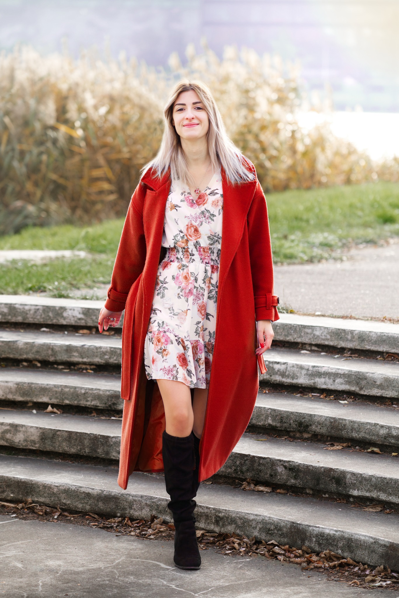 Uśmiechnięta kobieta w kwiecistej sukience i długim, pomarańczowym płaszczu schodzi po betonowych schodach na tle zarośli i wody.