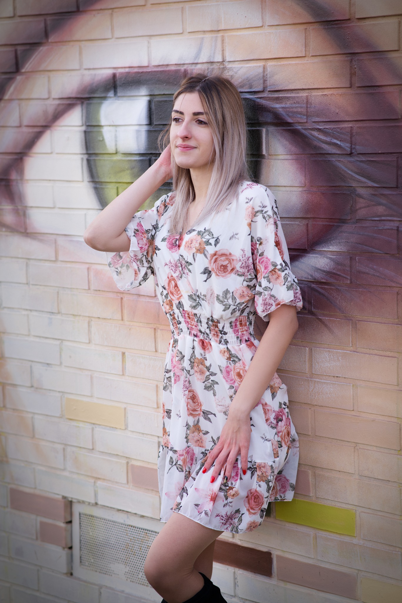 Kobieta w kwiecistej sukience pozuje na tle ceglanej ściany z muralem oka. Subtelny uśmiech, naturalne światło, swobodna poza. Sesja w plenerze, artystyczne tło.