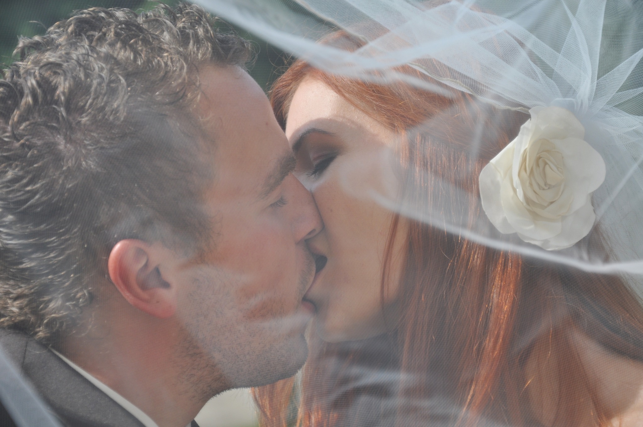 Nowożeńcy w pocałunku, widoczni przez welon z białą różą, zbliżenie na twarze.