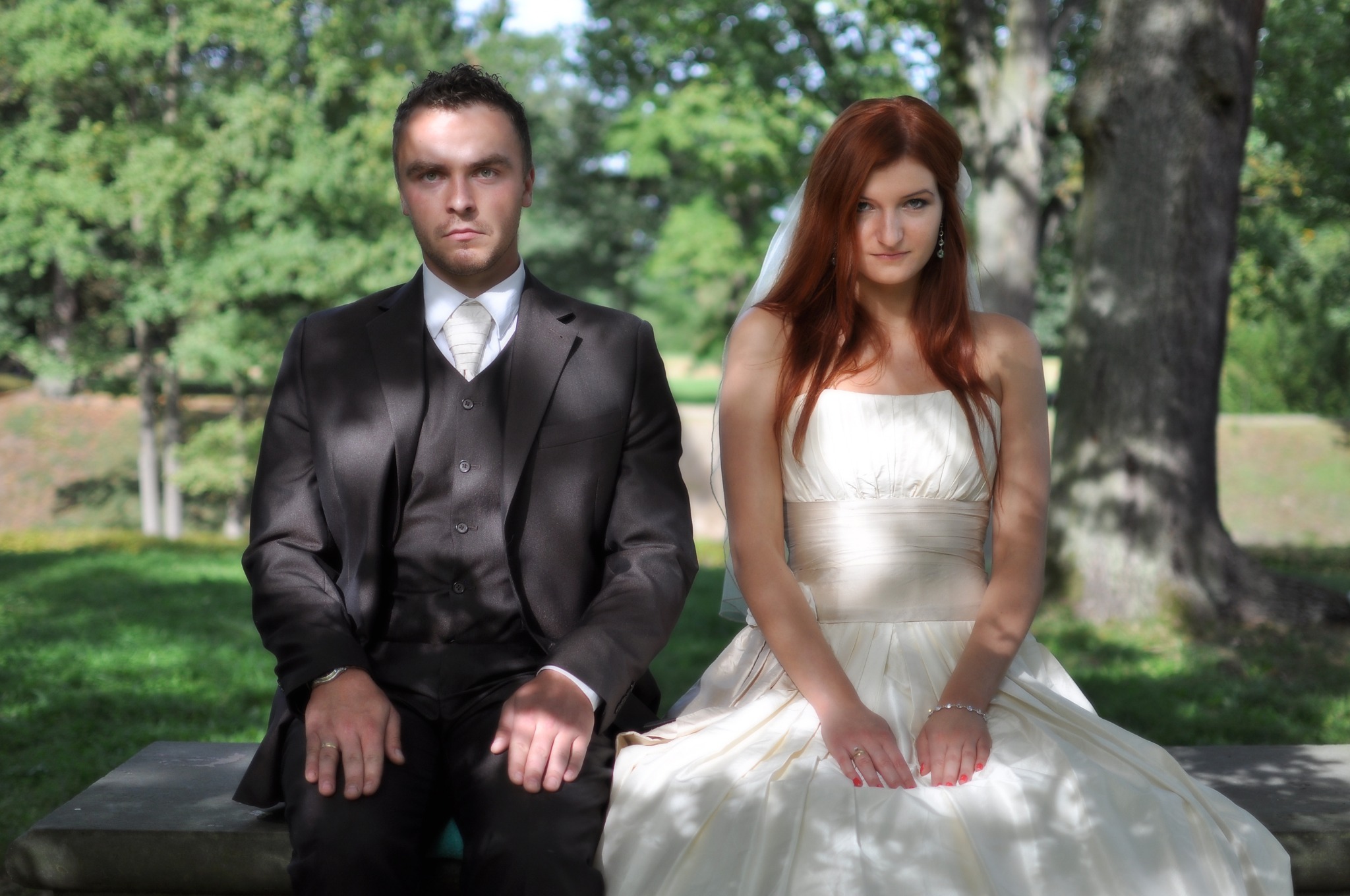 Para młoda siedząca na ławce w parku, on w ciemnym garniturze z kamizelką i jasnym krawacie, ona w białej sukni ślubnej z welonem i delikatną biżuterią, oboje patrzą w obiektyw z poważnym wyrazem...