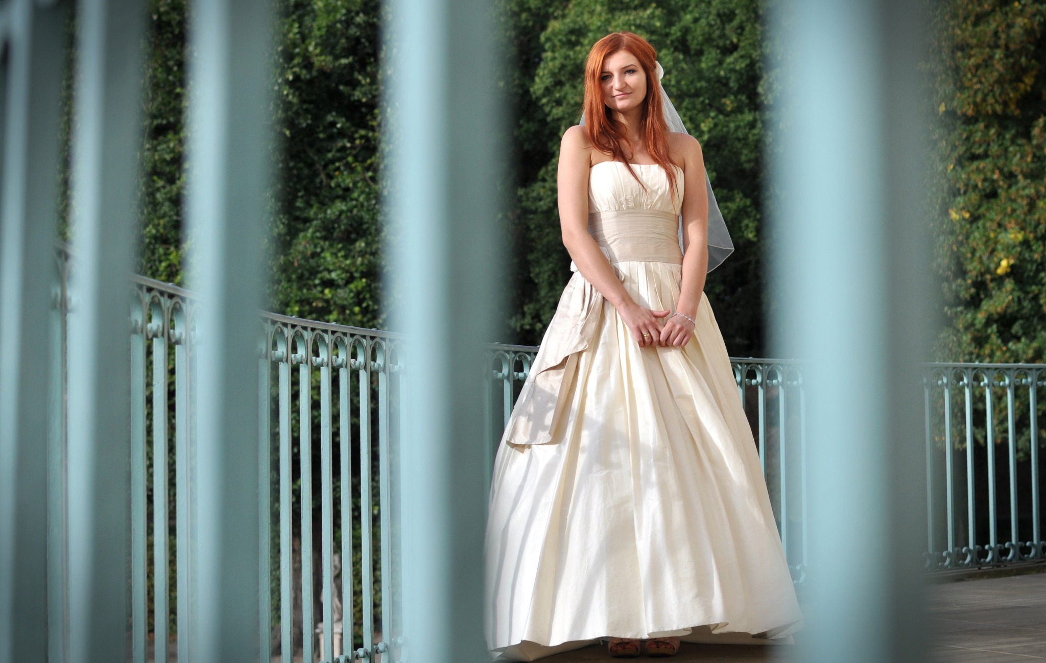 Panna młoda w kremowej sukni ślubnej z welonem, pozująca na balkonie z zielonym tłem, widoczna przez barierki.