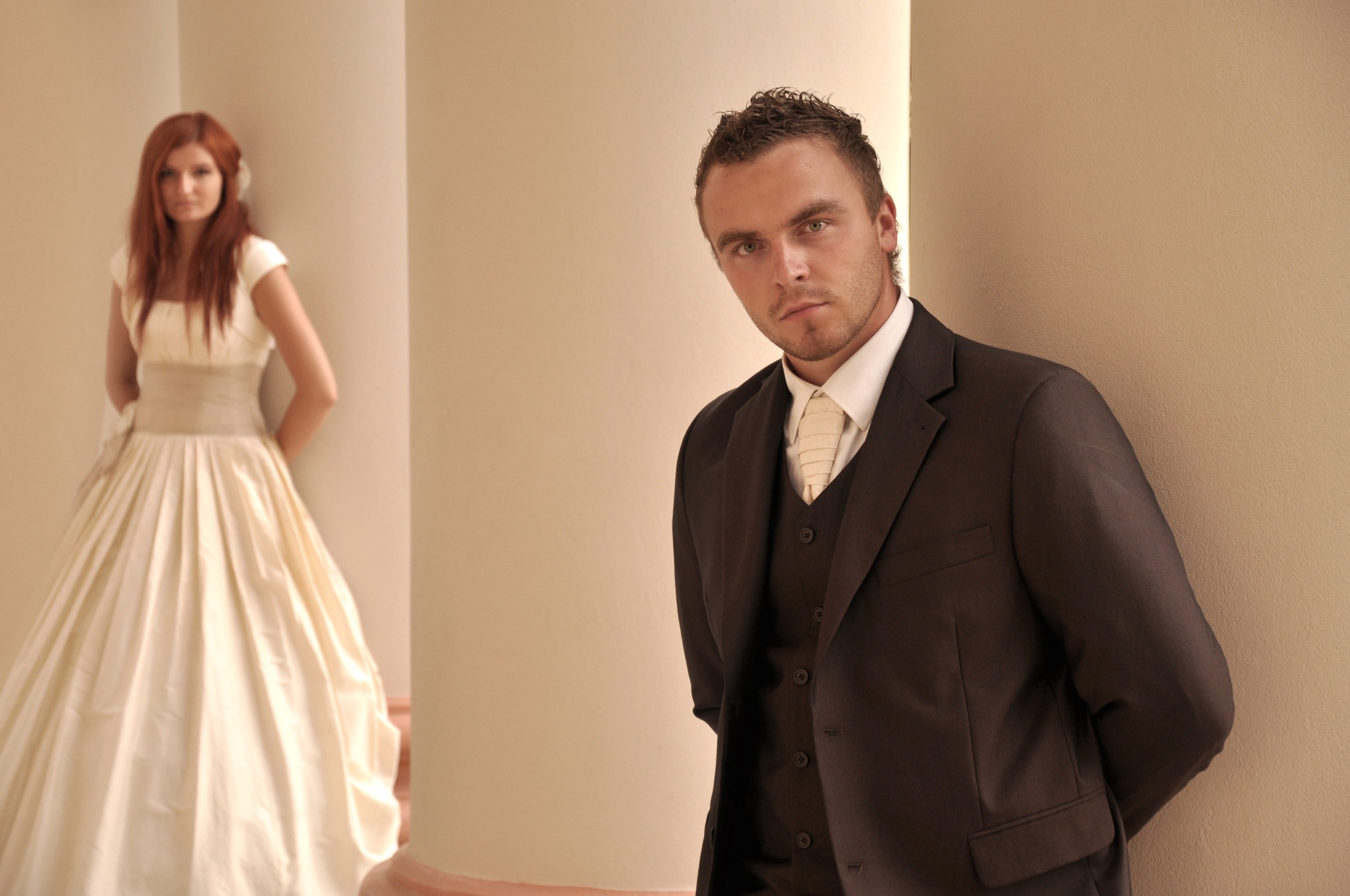 Elegancki pan w brązowym garniturze i kremowym krawacie, spoglądający w obiektyw, z rudowłosą kobietą w jasnej sukni balowej opartą o kolumnę w tle.