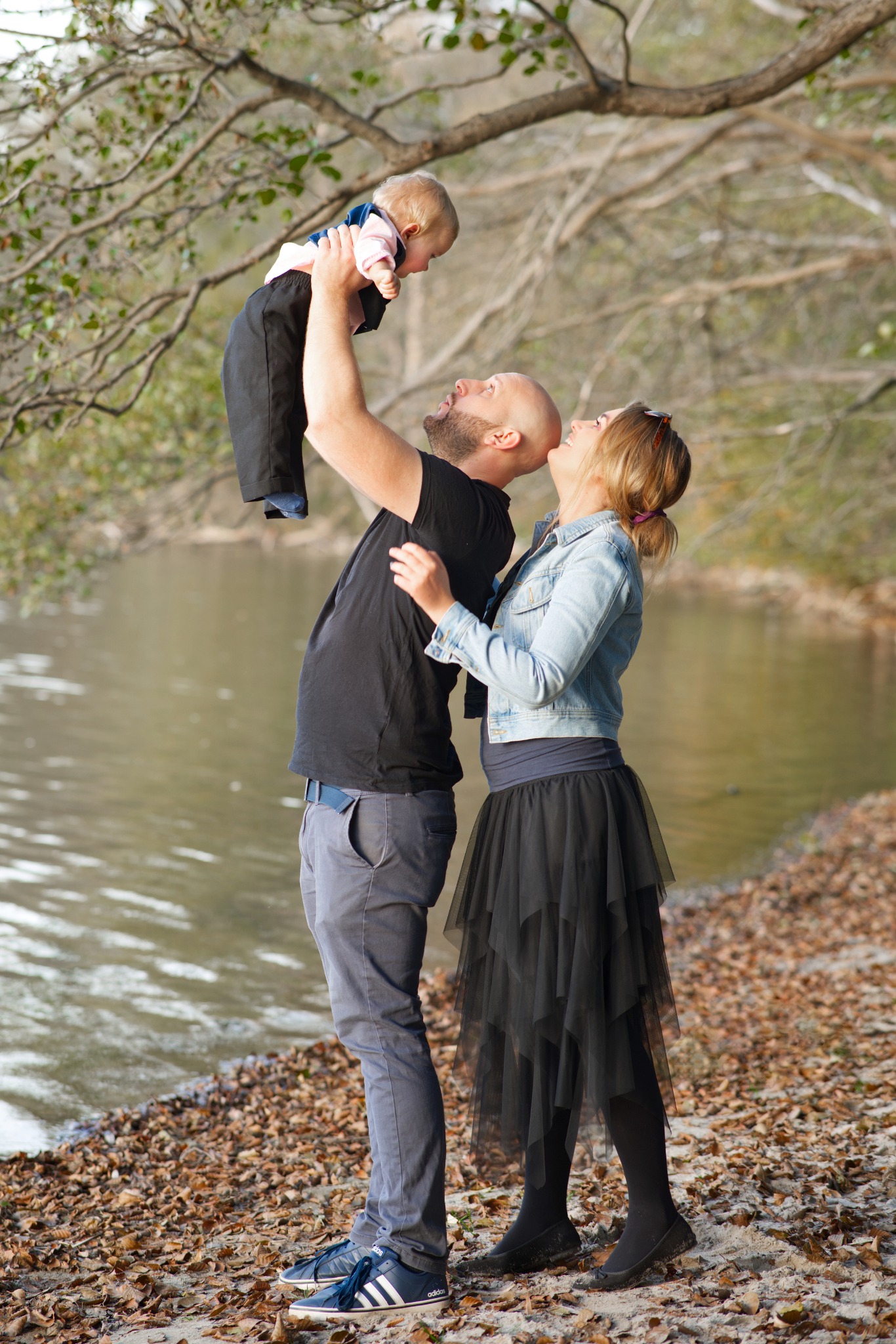 Rodzina nad brzegiem jeziora: ojciec podnosi w górę uśmiechnięte dziecko, matka z czułością patrzy na nich.