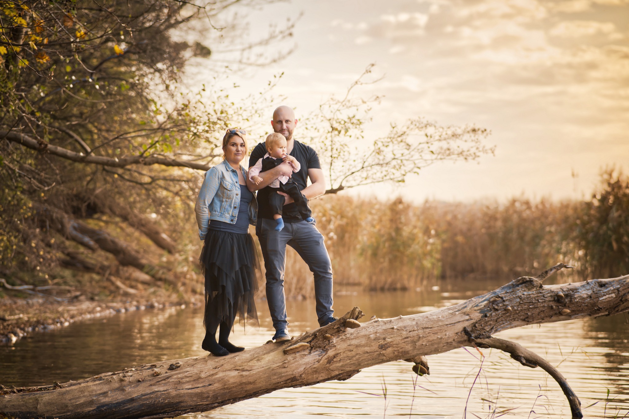 Rodzina z małym dzieckiem pozuje na powalonym drzewie nad wodą, w tle jesienny krajobraz.