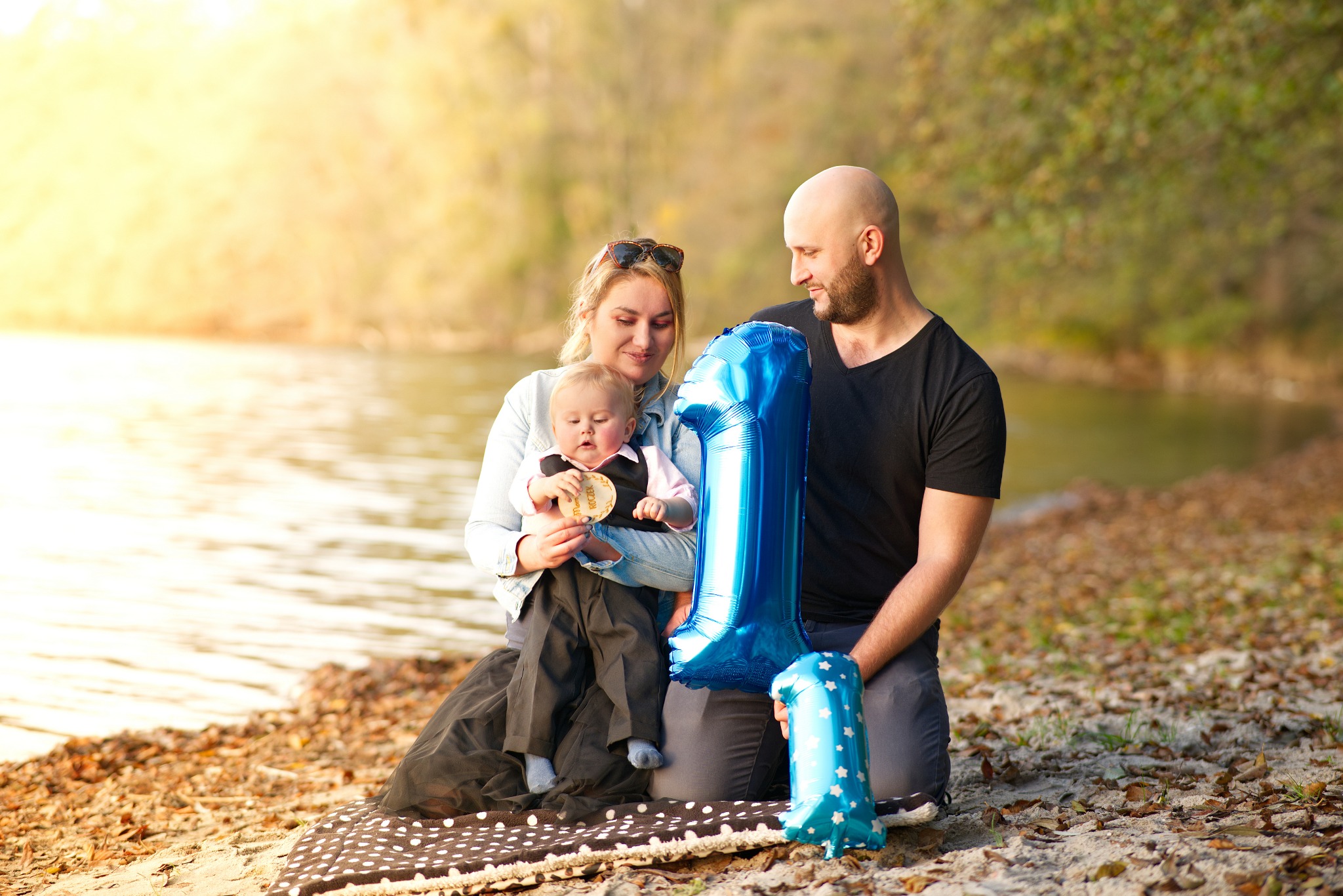 Rodzinne zdjęcie plenerowe: mama trzyma rocznego synka w ubranku wizytowym, tata obok z niebieskim balonem w kształcie cyfry 1, w tle woda i drzewa.