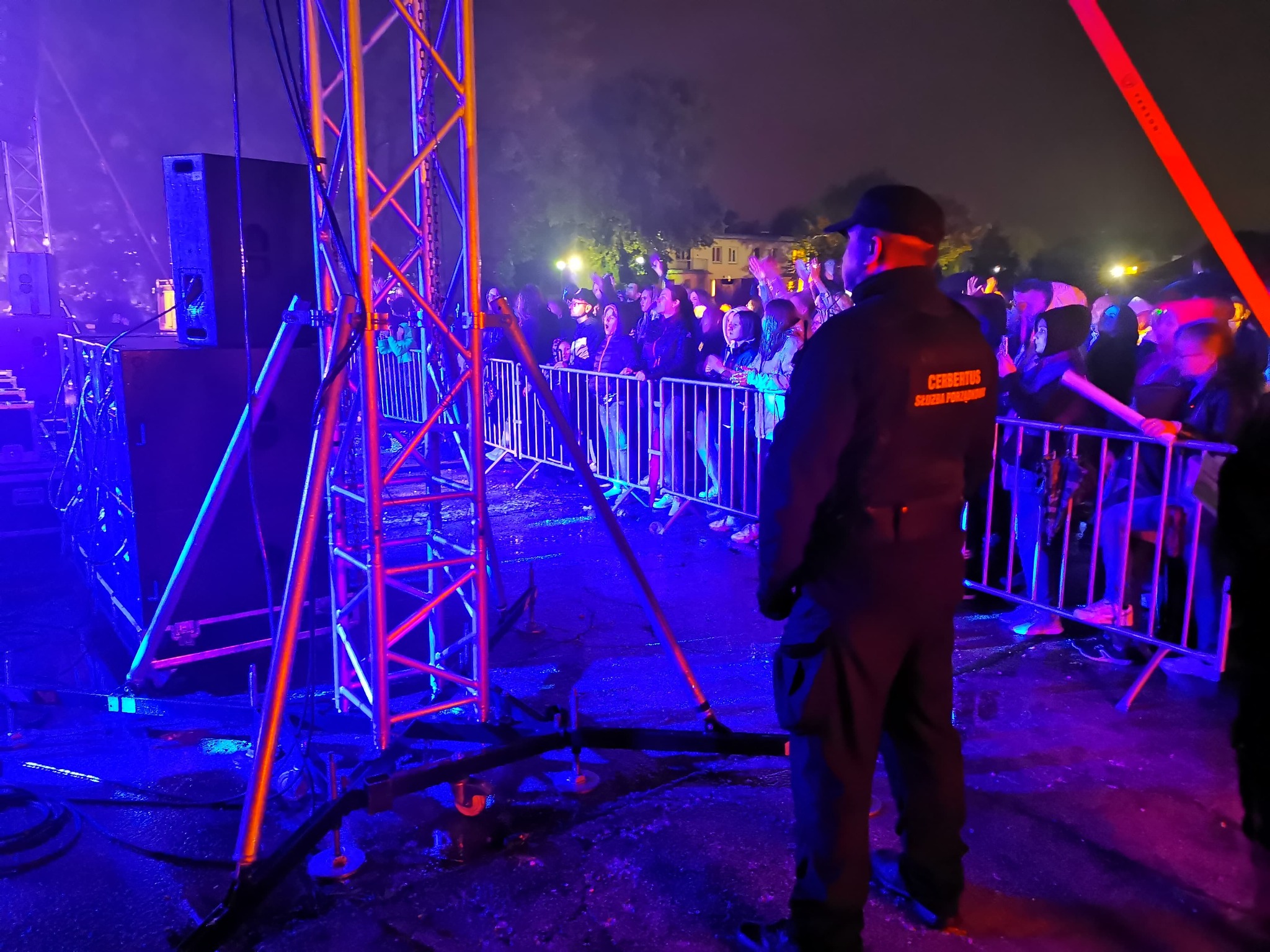 Ochroniarz w czarnym mundurze stoi tyłem na tle tłumu ludzi za metalową barierką, scena oświetlona niebieskim i pomarańczowym światłem.