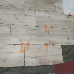 Układanie płytek imitujących drewno z użyciem klipsów poziomujących i krzyżyków dystansowych, widoczny fragment ściany z białymi płytkami i nałożony klej.