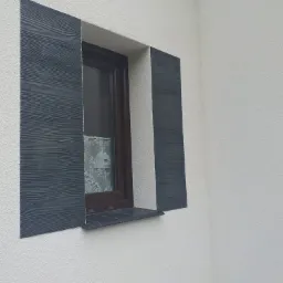 Wykończone okno z dekoracyjnymi panelami elewacyjnymi imitującymi drewno, zamontowanymi po bokach na białej, chropowatej elewacji.