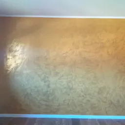 Ściana pomalowana dekoracyjną farbą w odcieniach złota i beżu z widoczną fakturą, oświetlona z lewej strony, z drewnianą podłogą i rośliną doniczkową na drewnianym stoliku.