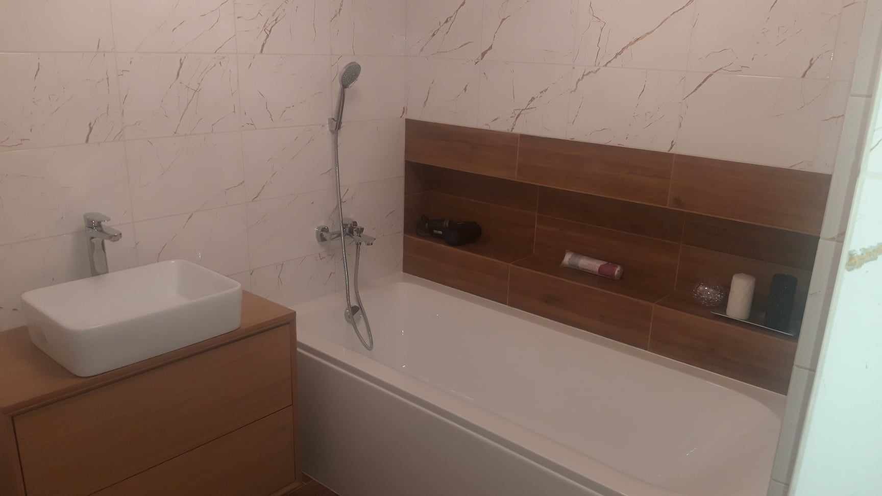 Wykończona łazienka z białymi marmurkowymi płytkami, drewnianą szafką pod umywalką, wnęką nad wanną wyłożoną drewnopodobnymi płytkami i chromowaną armaturą.