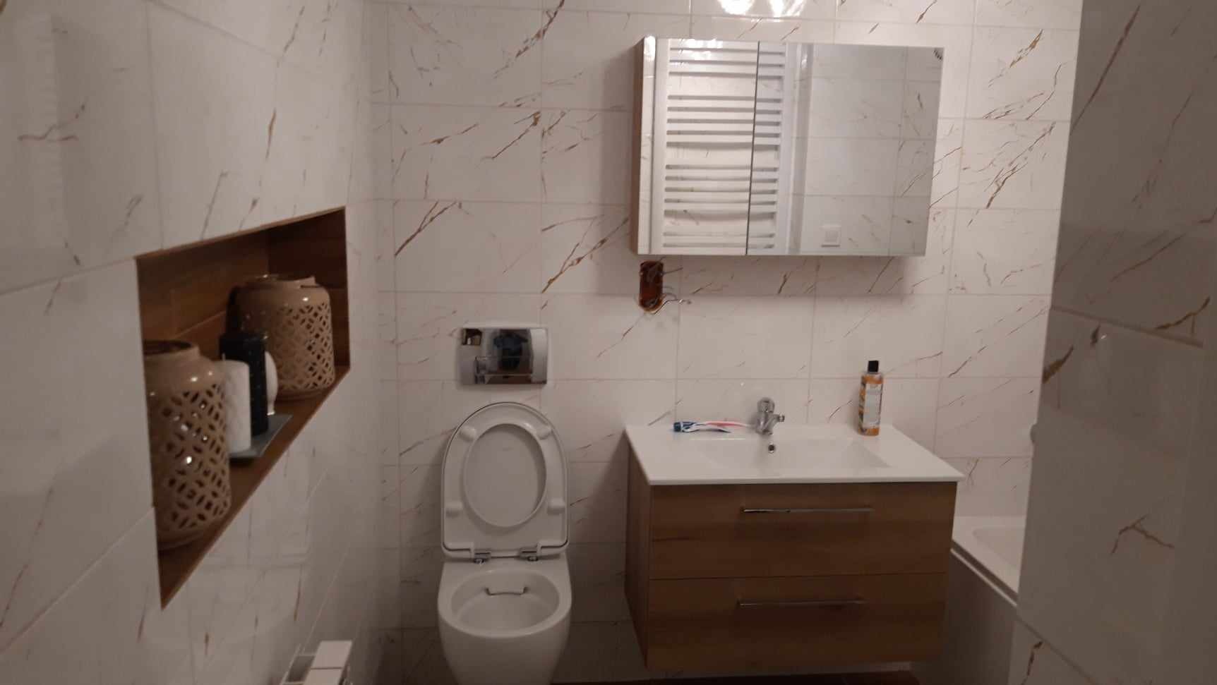 Nowoczesna łazienka z białymi płytkami imitującymi marmur, podwieszana toaleta z otwartą klapą, szafka pod umywalką z drewnianymi frontami, lustro z szafką nad umywalką, wnęka w ścianie...