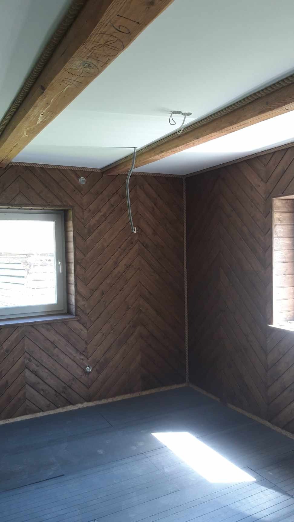 Wykończone wnętrze pokoju z drewnianą boazerią ułożoną w jodełkę na ścianach, drewnianymi belkami stropowymi z dekoracyjnym sznurem, świeżo pomalowanym sufitem na biało i przygotowaną podłogą.