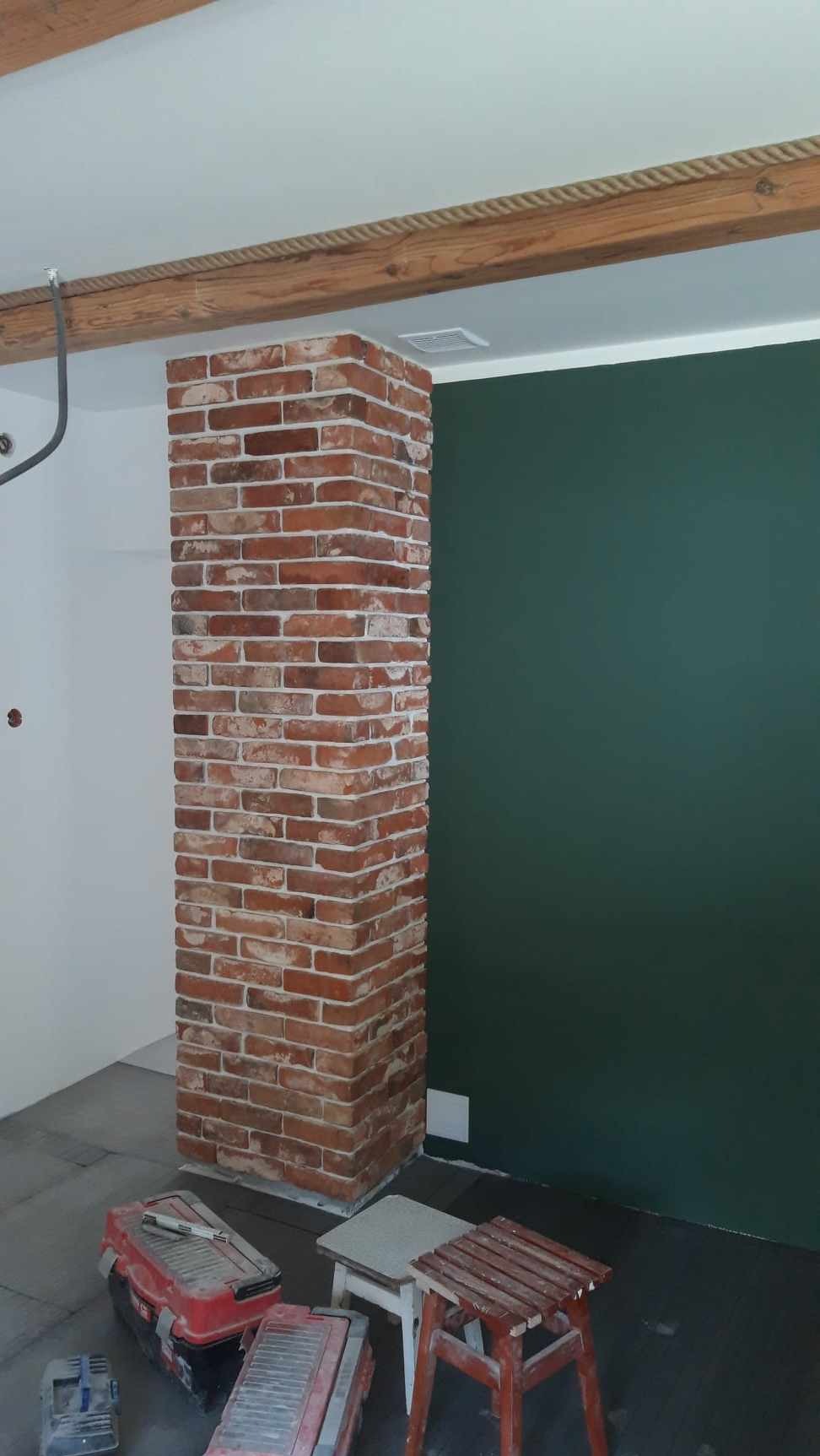 Wnętrze z kolumną obłożoną czerwoną cegłą, widoczne narzędzia i stołki robocze, ściana w kolorze butelkowej zieleni, drewniany belka stropowa z dekoracyjnym sznurem.