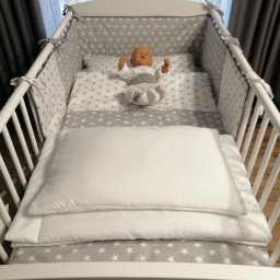 Luli Laj - Białe łóżeczko dziecięce z szarym ochraniaczem i pościelą w białe gwiazdki, z manekinem dziecka w środku.