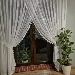 Delikatne, białe firany z koronkowym wykończeniem, upięte po bokach okna, z widocznym kwiatem doniczkowym w tle i na parapecie, na tle drewnianej podłogi.