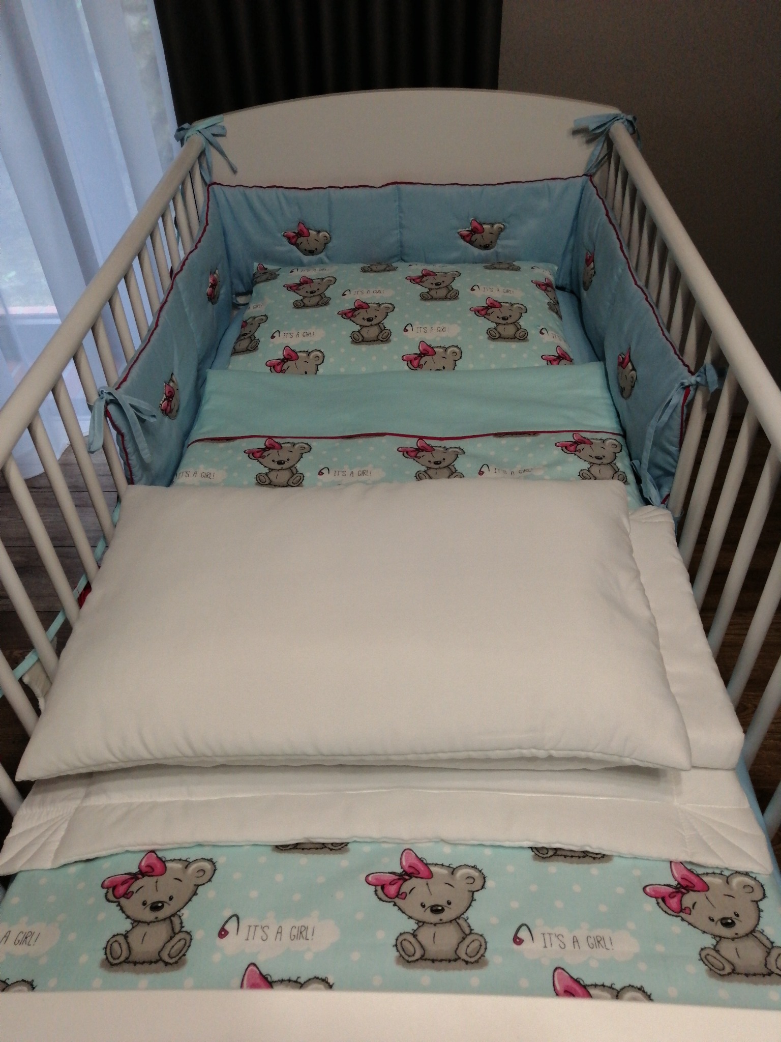 Białe łóżeczko dziecięce z błękitną pościelą w misie z różowymi kokardkami i napisem 'It's a girl!'