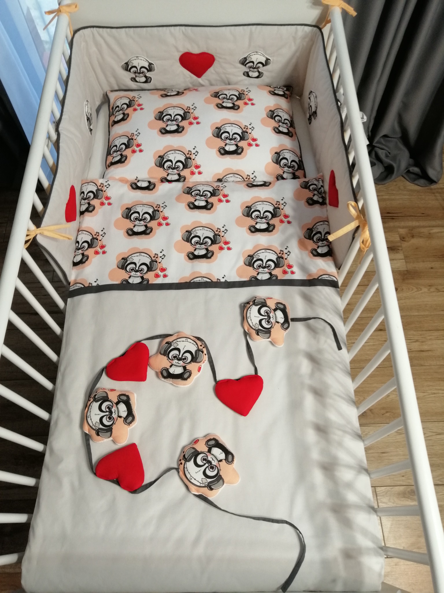 Wyposażenie dziecięcego łóżeczka: pościel z motywem pand w słuchawkach i czerwone serduszka, widok z góry na białe łóżeczko z drewnianą podłogą w tle.