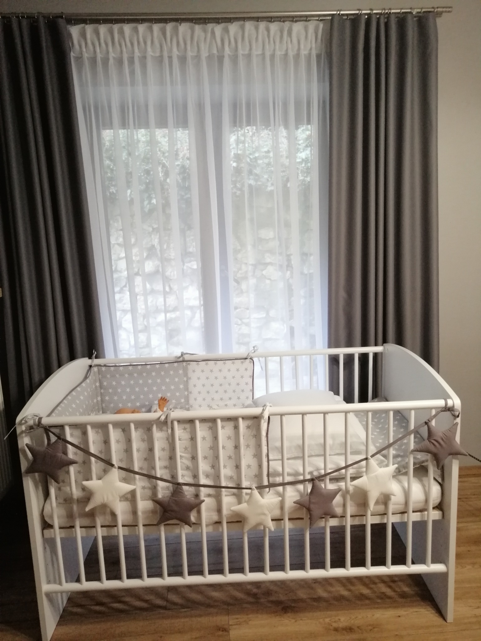 Białe łóżeczko dziecięce z szarym ochraniaczem w gwiazdki i girlandą z gwiazd na tle okna z szarymi i białymi zasłonami.