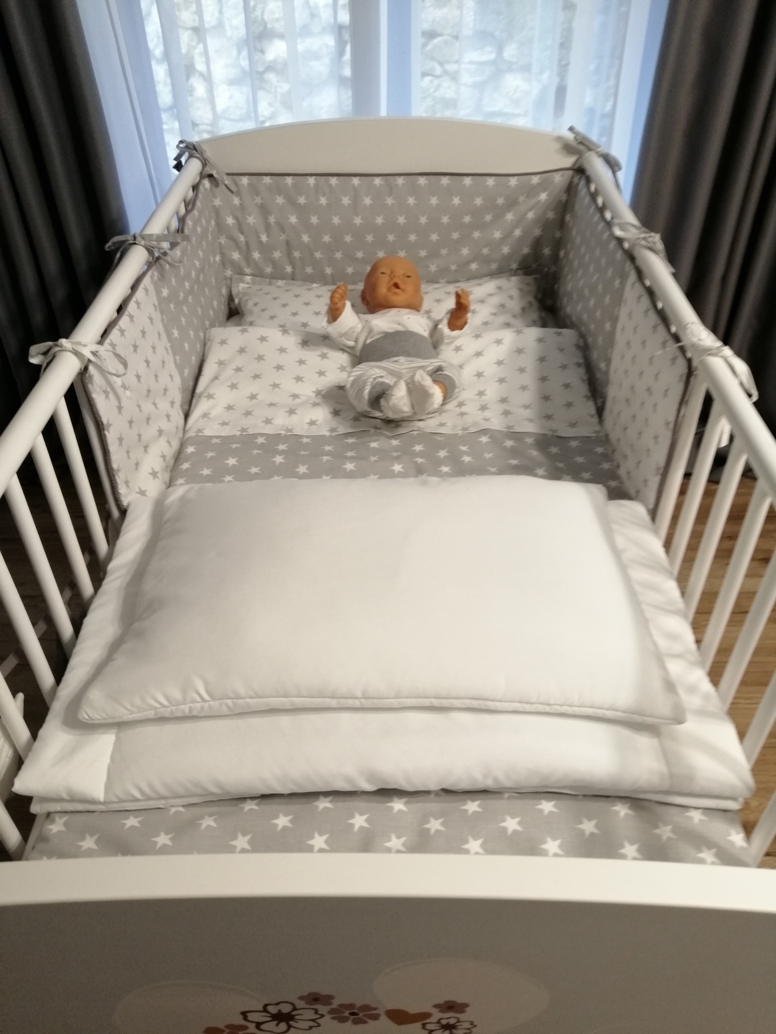 Białe łóżeczko dziecięce z szarym ochraniaczem i pościelą w białe gwiazdki, z manekinem dziecka w środku.