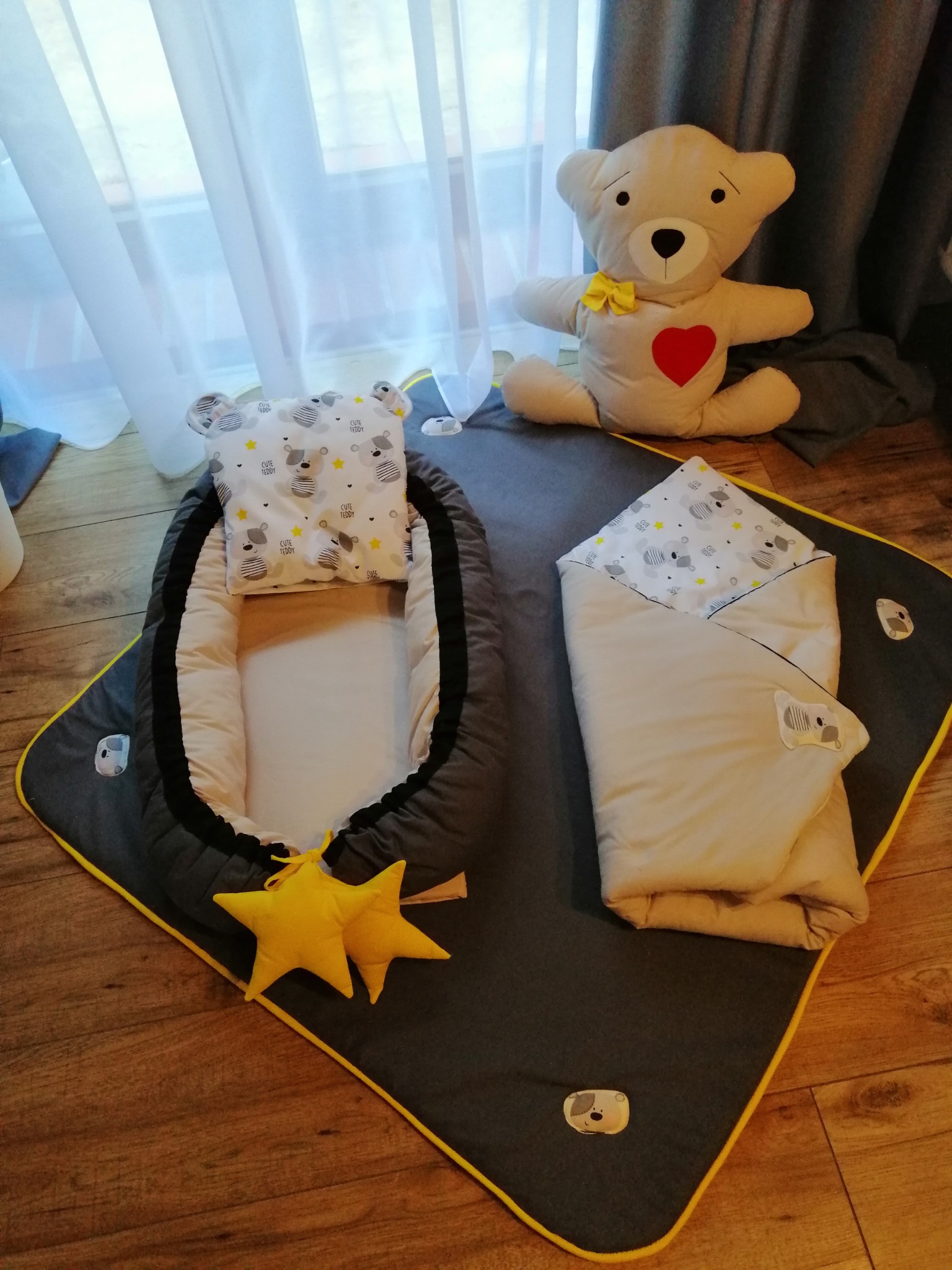 Zestaw niemowlęcy: kokon, poduszka z uszkami, rożek i mata do zabawy w odcieniach szarości, beżu i żółci, z motywem misiów i gwiazdek, pluszowy miś w tle.