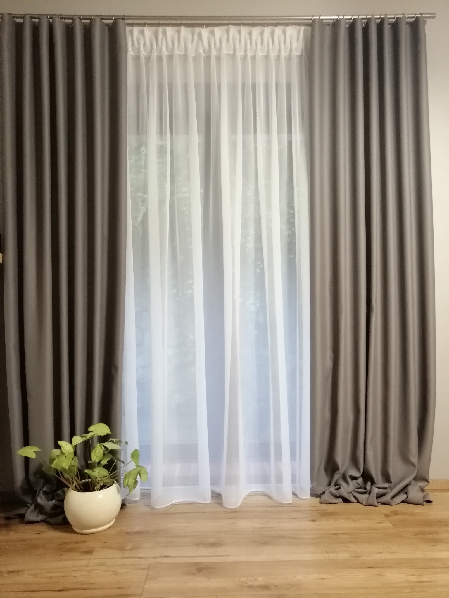 Zasłony zaciemniające w odcieniach szarości i firanka woalowa w oknie, z rośliną doniczkową w kremowej osłonce na podłodze.
