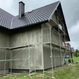 Dom jednorodzinny w trakcie tynkowania elewacji, widoczne rusztowania i świeżo nałożony tynk w szarym odcieniu, dach pokryty ciemną dachówką.
