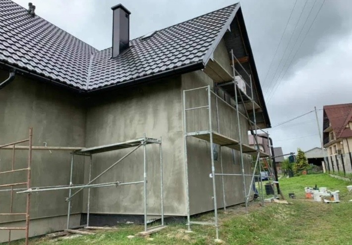 Dom jednorodzinny w trakcie tynkowania elewacji, widoczne rusztowania i świeżo nałożony tynk w szarym odcieniu, dach pokryty ciemną dachówką.