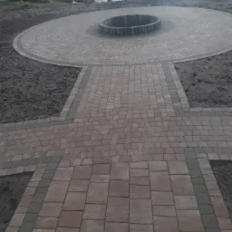 Okrągły plac z kostki brukowej z centralnym paleniskiem, połączony ścieżką z kostki, widok na staw i budynek w tle.