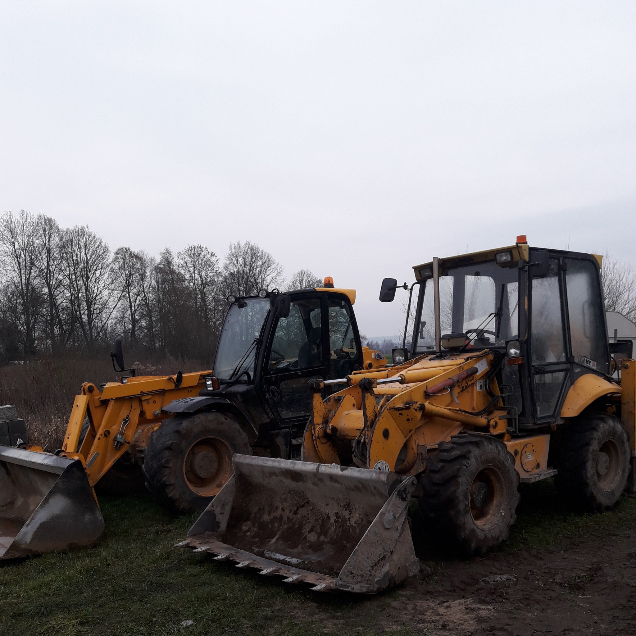 Dwa żółto-czarne traktory z łyżkami stojące na ziemi w Ornecie, w tle drzewa bez liści i pochmurne niebo.