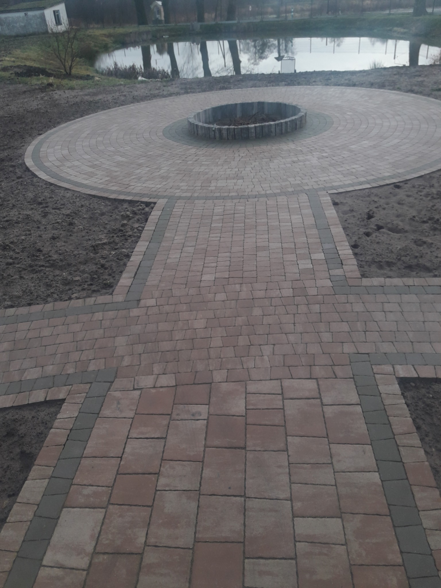 Okrągły plac z kostki brukowej z centralnym paleniskiem, połączony ścieżką z kostki, widok na staw i budynek w tle.