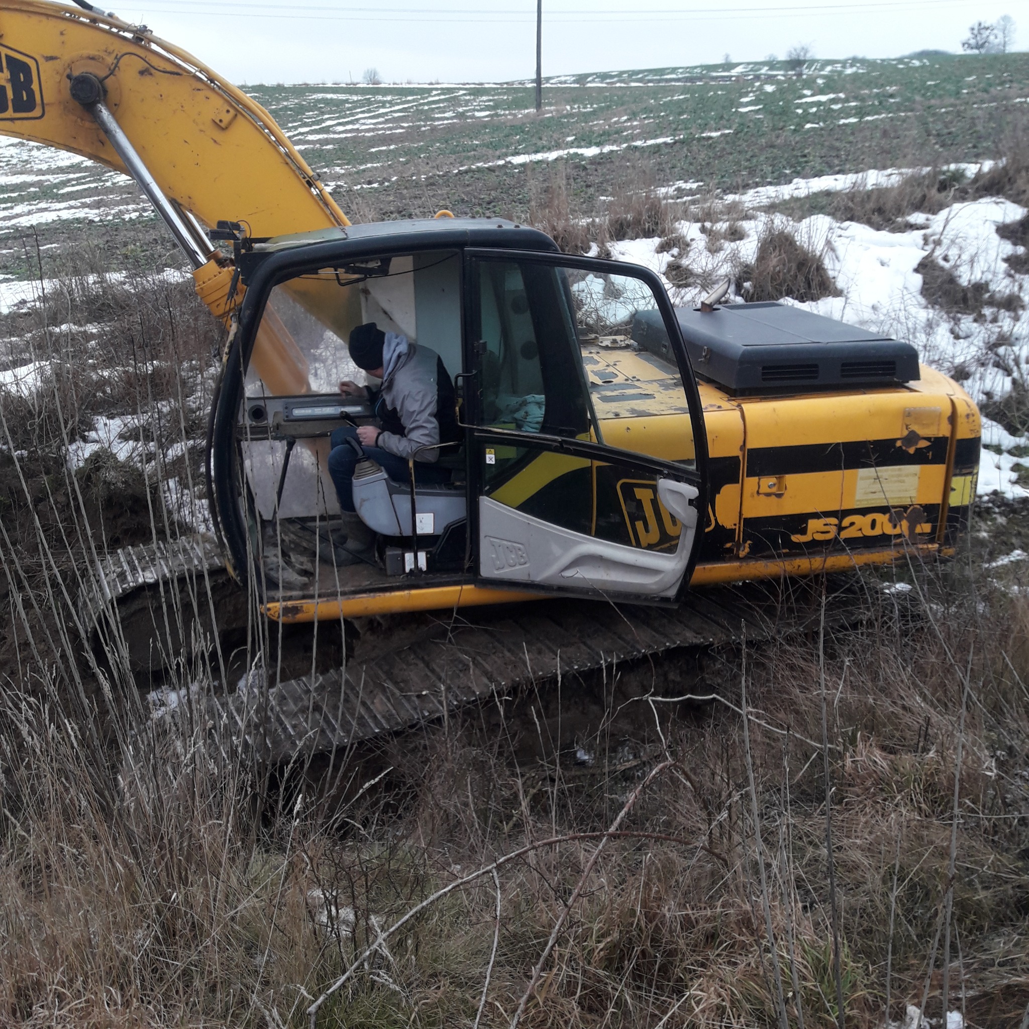 Żółta koparka JCB JS200 z otwartymi drzwiami kabiny, operator w środku, teren za koparką częściowo pokryty śniegiem, widoczne suche trawy na pierwszym planie.