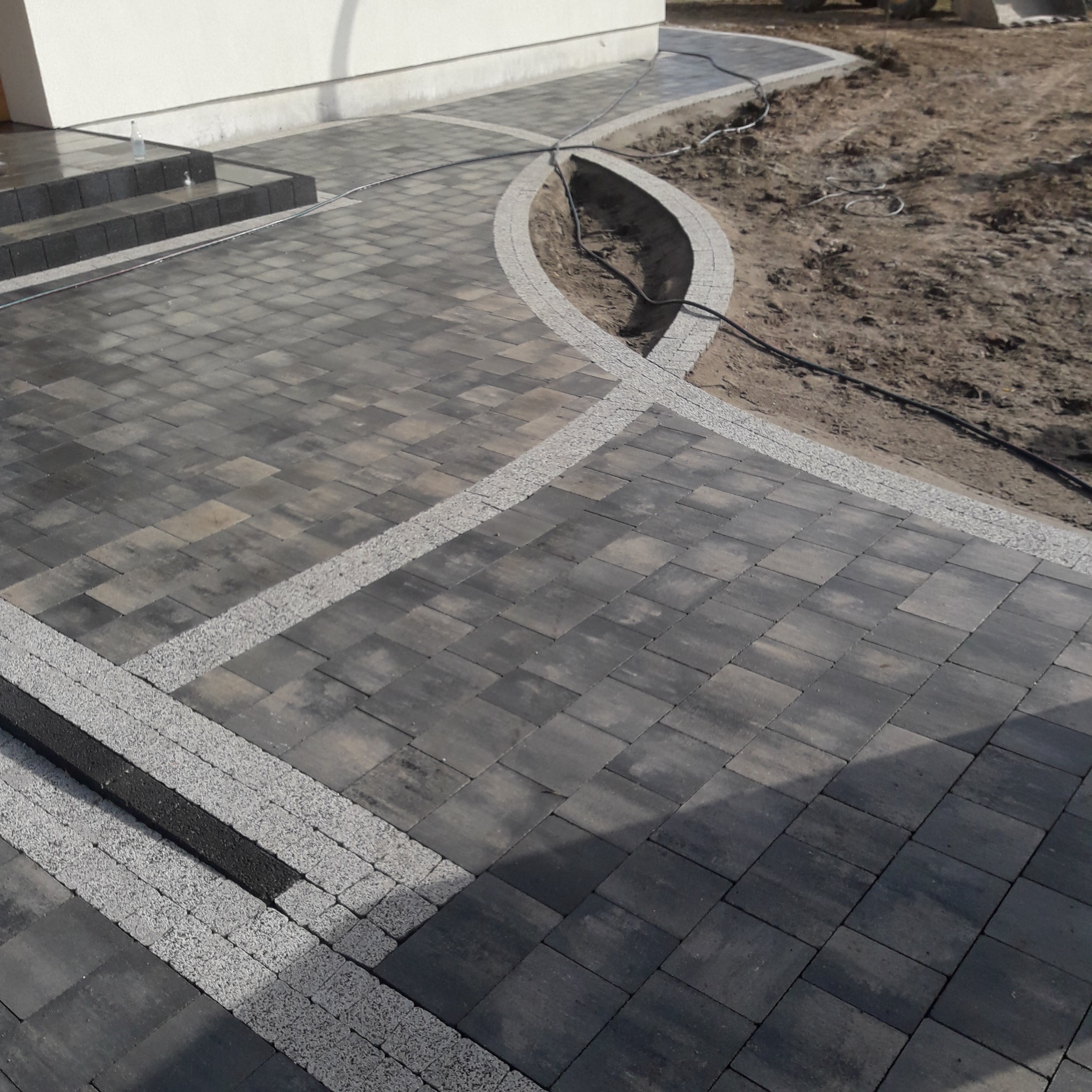 Ułożona, ciemnoszara kostka brukowa z jasnoszarą obwódką, tworząca ścieżkę i schody przed domem. Widoczny fragment rabaty z ziemią. Na schodach stoi butelka z wodą.