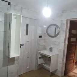 Wykończona łazienka z szarymi płytkami imitującymi kamień, kabiną prysznicową z czarnymi profilami, wiszącą szafką, umywalką z szafką oraz okrągłym lustrem