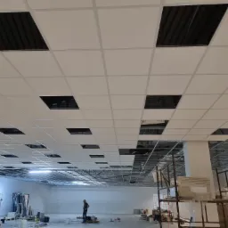 Widok z dołu na biały sufit podwieszany kasetonowy z czarnymi otworami na oświetlenie, widoczne elementy konstrukcyjne nad sufitem, w tle osoba pracująca i rusztowania.