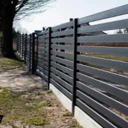 Nowoczesne ogrodzenie z poziomych, metalowych profili w kolorze grafitowym, osadzone na betonowej podmurówce, widok z perspektywy bocznej na tle drzew i zabudowań.