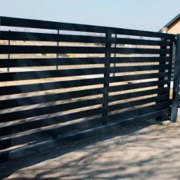 Nowoczesna, pozioma brama wjazdowa i przyległe ogrodzenie z metalowych profili pomalowanych na grafitowo, z widocznym domem w tle.